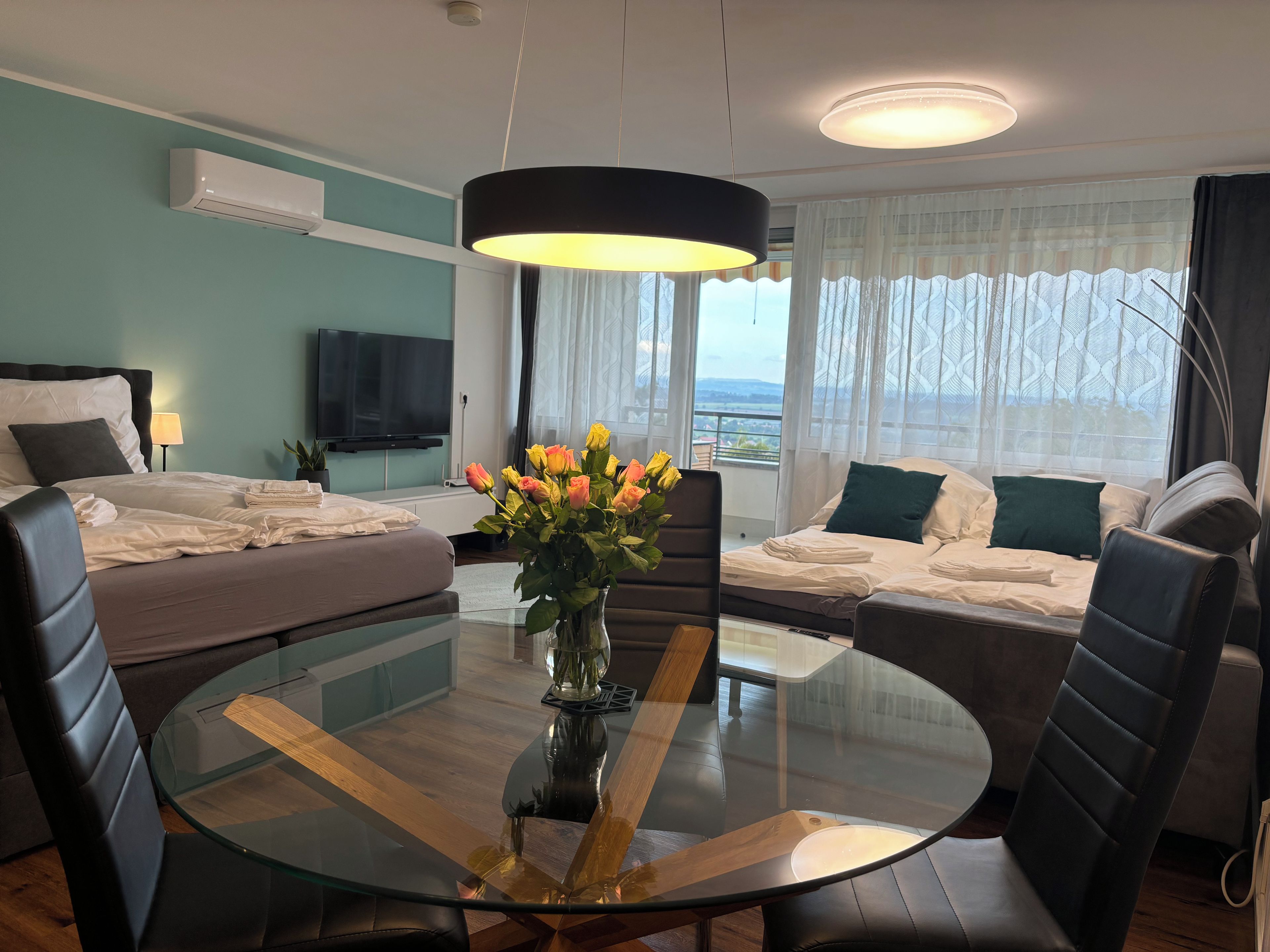 Quarto de hotel moderno com mesa de jantar de vidro, dois sofás, televisão e varanda com vista.