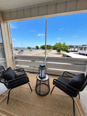 A balcony with two black wicker chairs and a table, overlooking a street with parked cars and a body of water. - Spacious 2BR Near NASA + Pool (Houston)