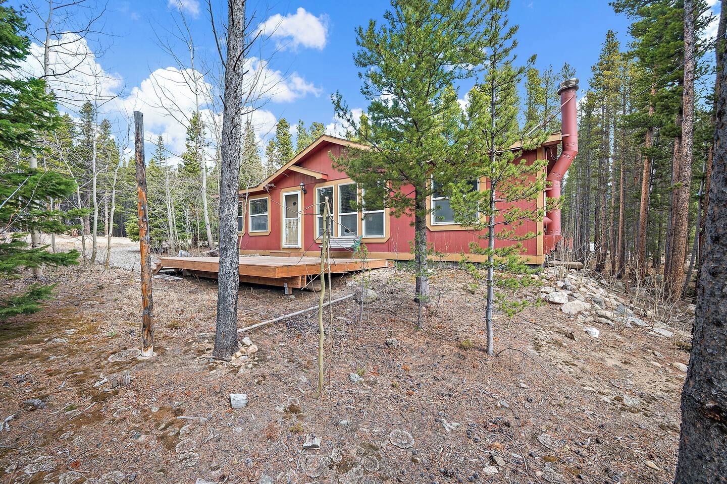A cabin with a wooden deck, surrounded by a forest with tall pine trees.