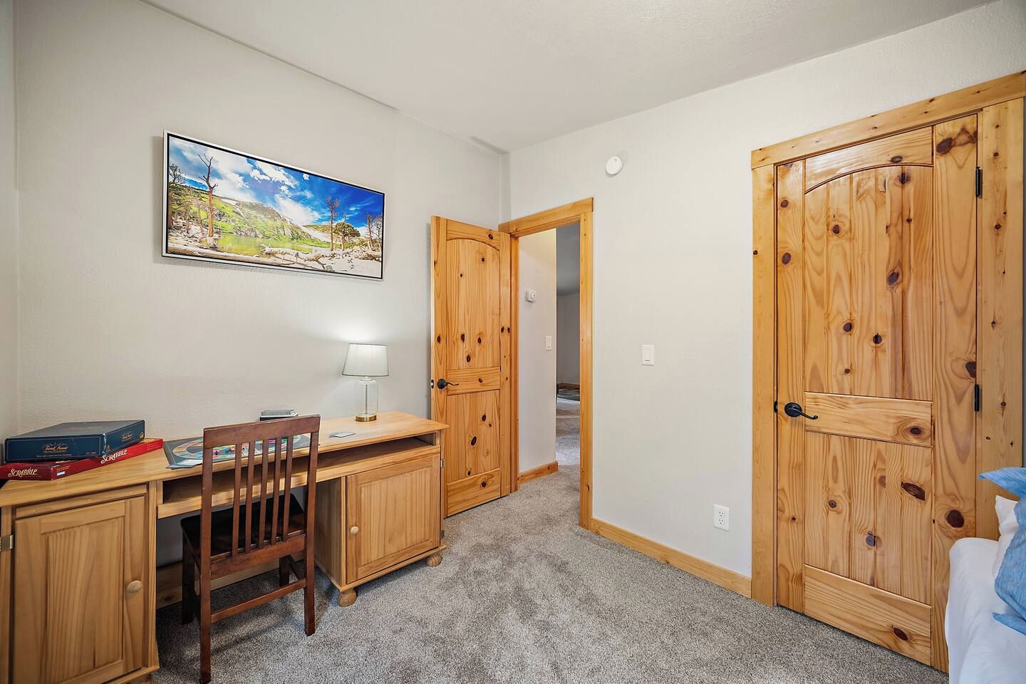 A room with a desk, chair, and a painting on the wall. There are two wooden doors and a carpeted floor.