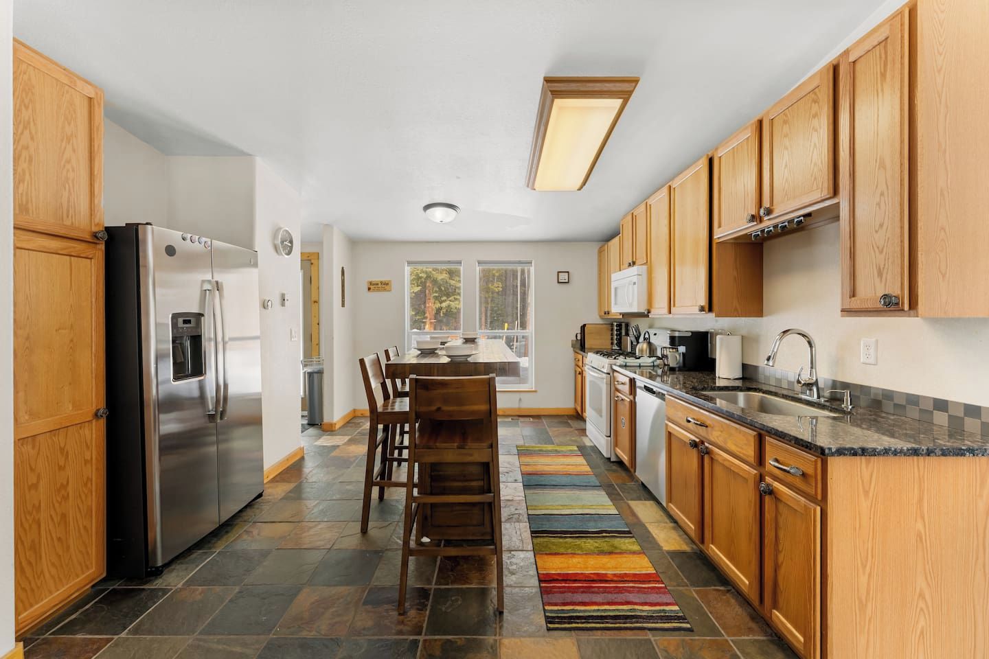 A modern kitchen with a stainless steel refrigerator, wooden cabinets, a granite countertop, and a colorful rug.