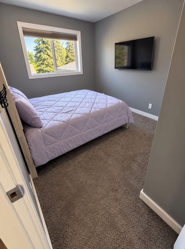 A bedroom with a bed, a window, and a flat-screen TV mounted on the wall.