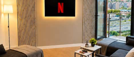 A modern living room with a large flat-screen TV mounted on the wall, a sofa set, a small table with a plant, and a view of the city through large windows.