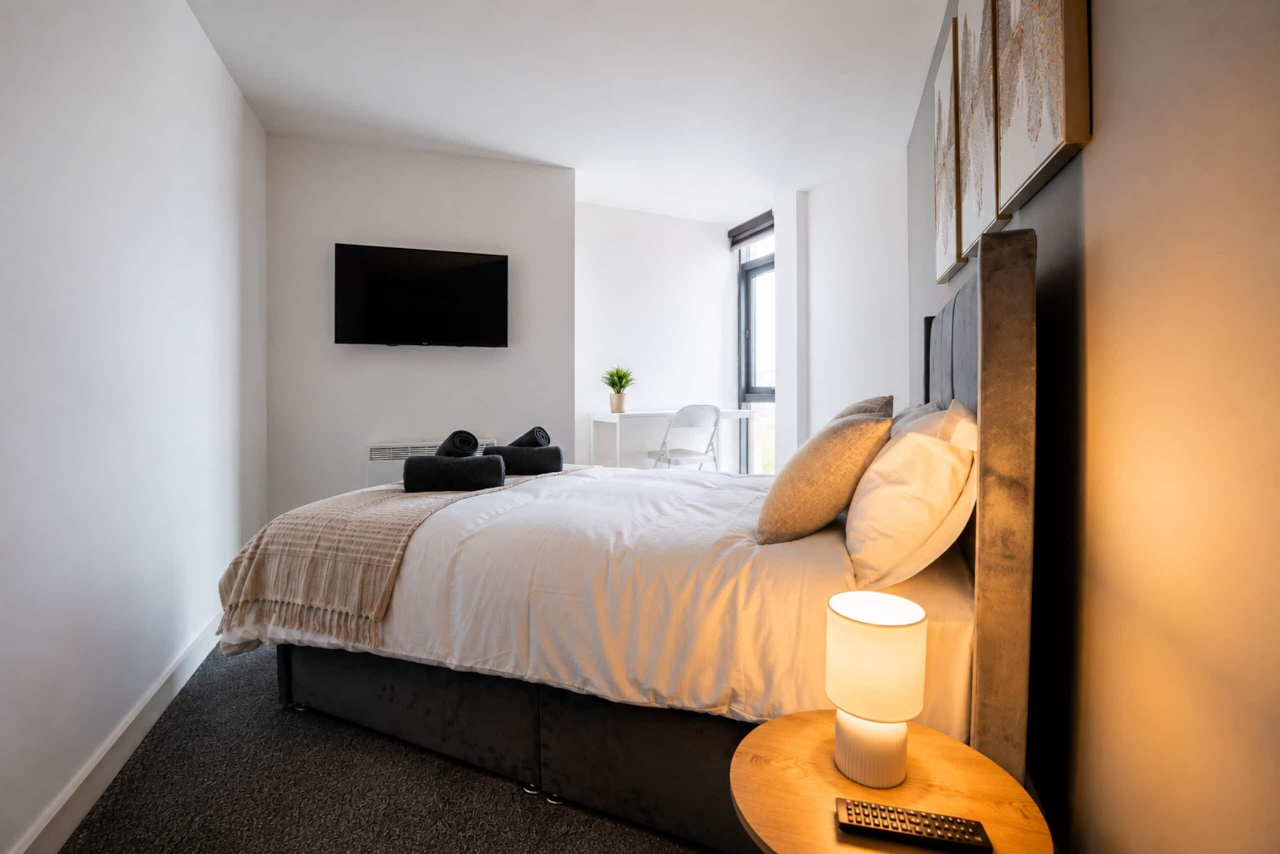 Ein modernes Hotelzimmer mit einem Bett, einer Nachttischlampe, einer Fernbedienung und einem an der Wand befestigten Flachbildfernseher.