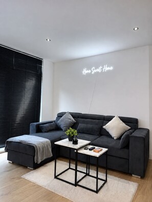 A modern living room with a dark grey sofa, a small side table, and a wall-mounted neon sign that reads 'Home Sweet Home.' - Modern Canal-Side 2BR - Balcony - Sleeps 8 (leeds)