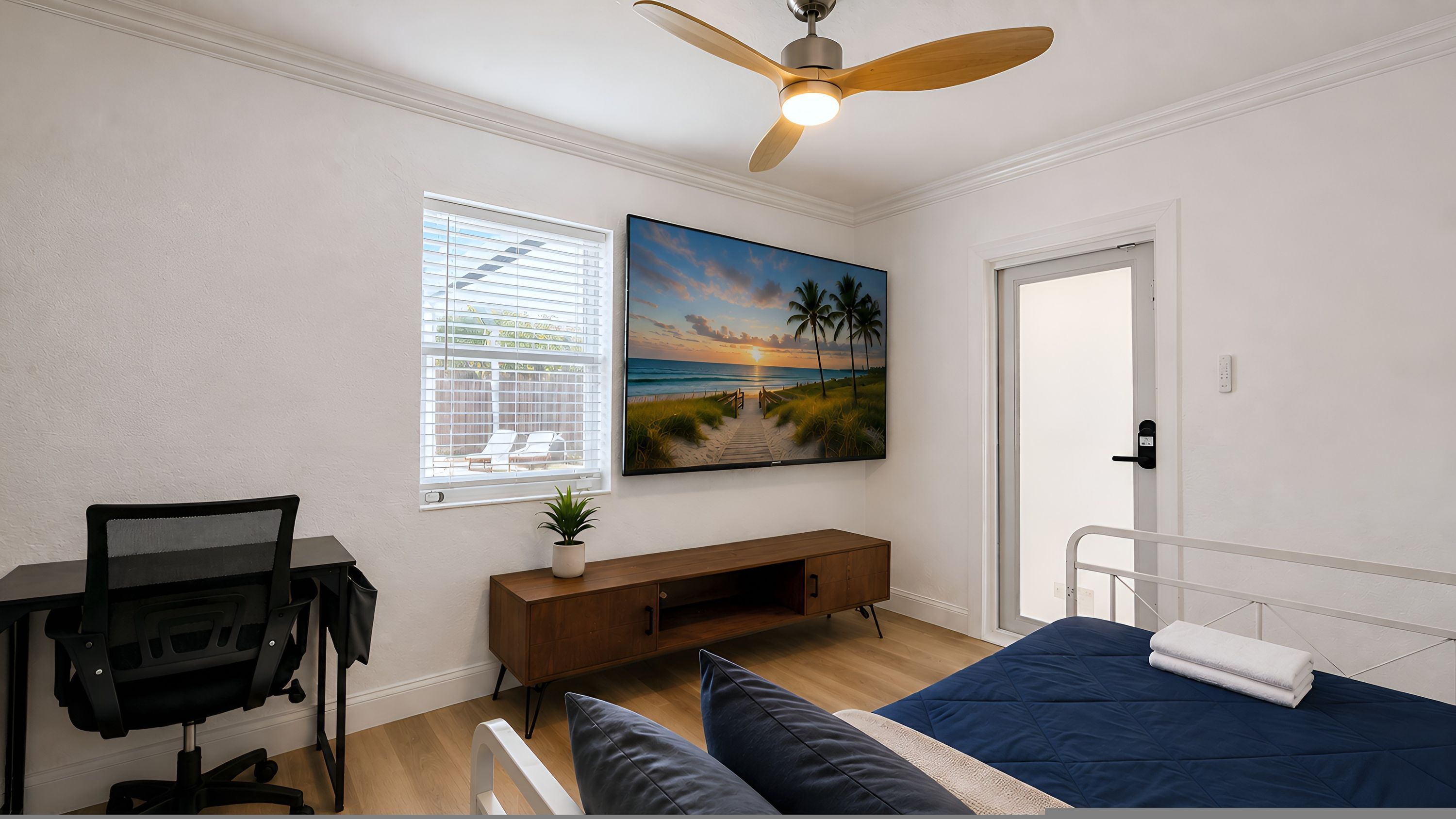 A bedroom with a bed, a desk, a chair, a ceiling fan, a window with blinds, and a painting of a beach scene.