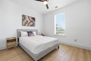 Et soveværelse med seng, sengebord, ventilator i loftet, et vindue og et kunstværk på væggen.