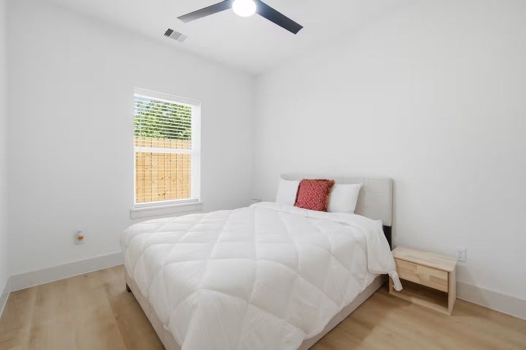 A bedroom with a bed, a nightstand, a window with blinds, and a ceiling fan.