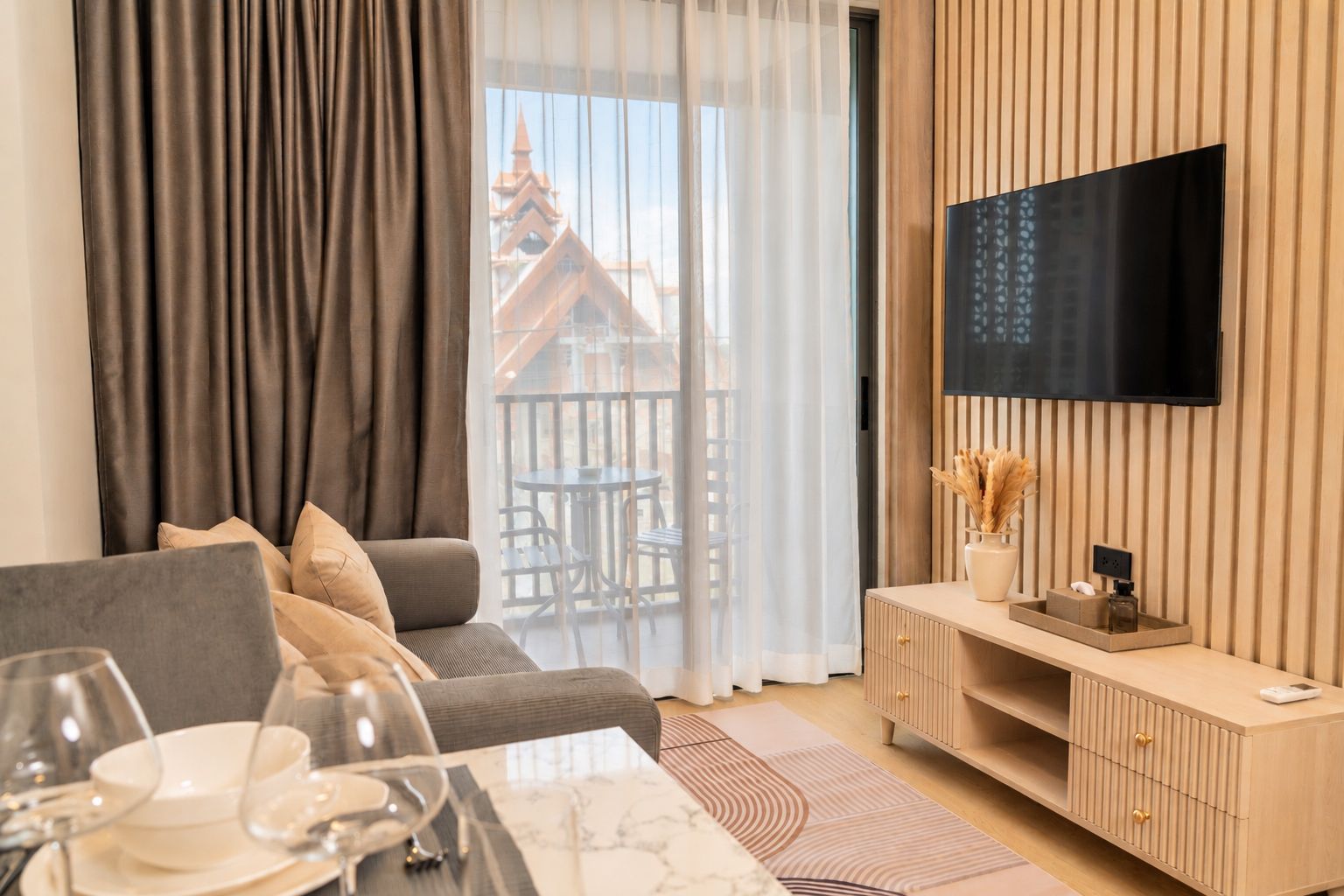 A modern living room with a sofa, dining table, and a flat-screen TV mounted on a wooden wall.
