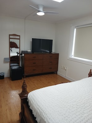 Un dormitorio con cama, cómoda, televisor, ventilador de techo y ventana.