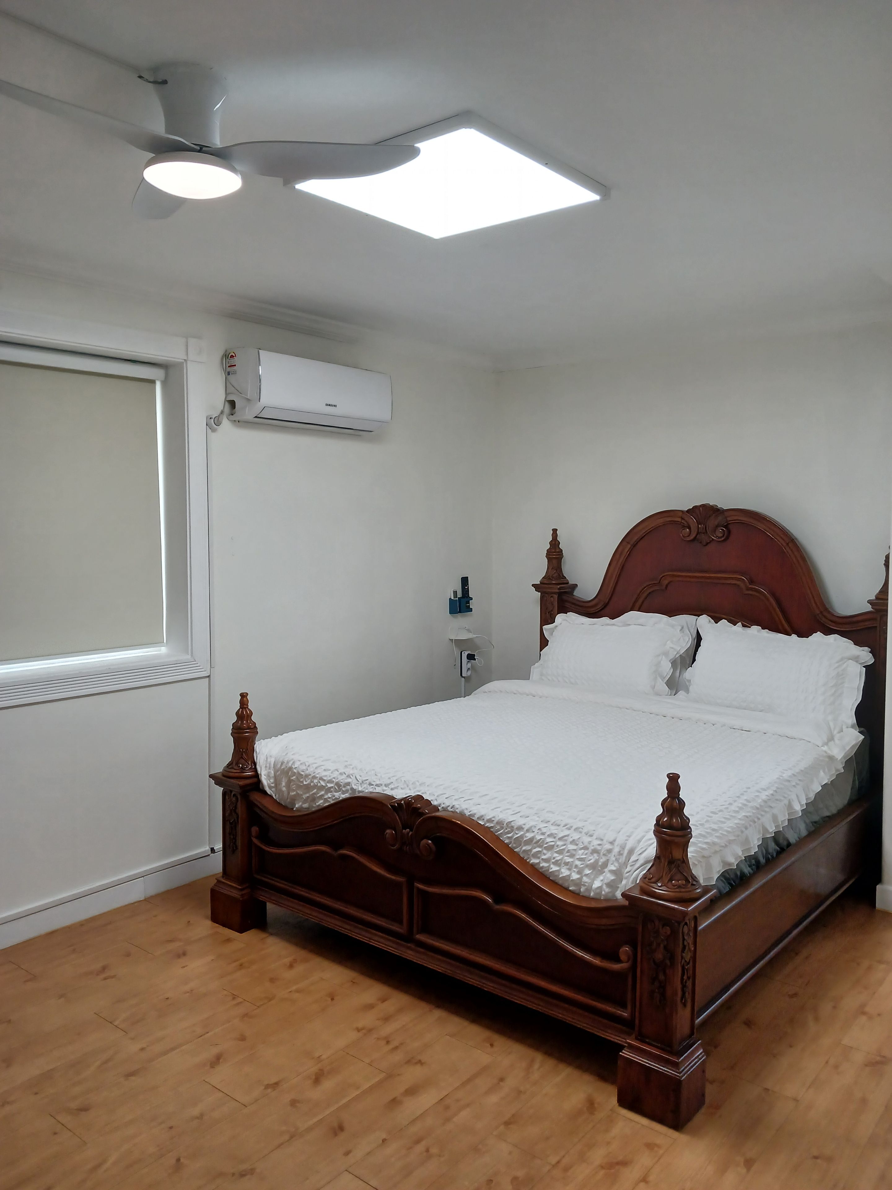 A bedroom with a wooden bed, white bedding, a ceiling fan, an air conditioning unit, and a window with blinds.
