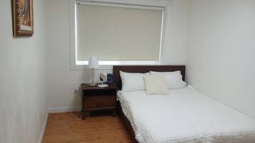 A bedroom with a bed, a nightstand, a lamp, a fan, and a window.