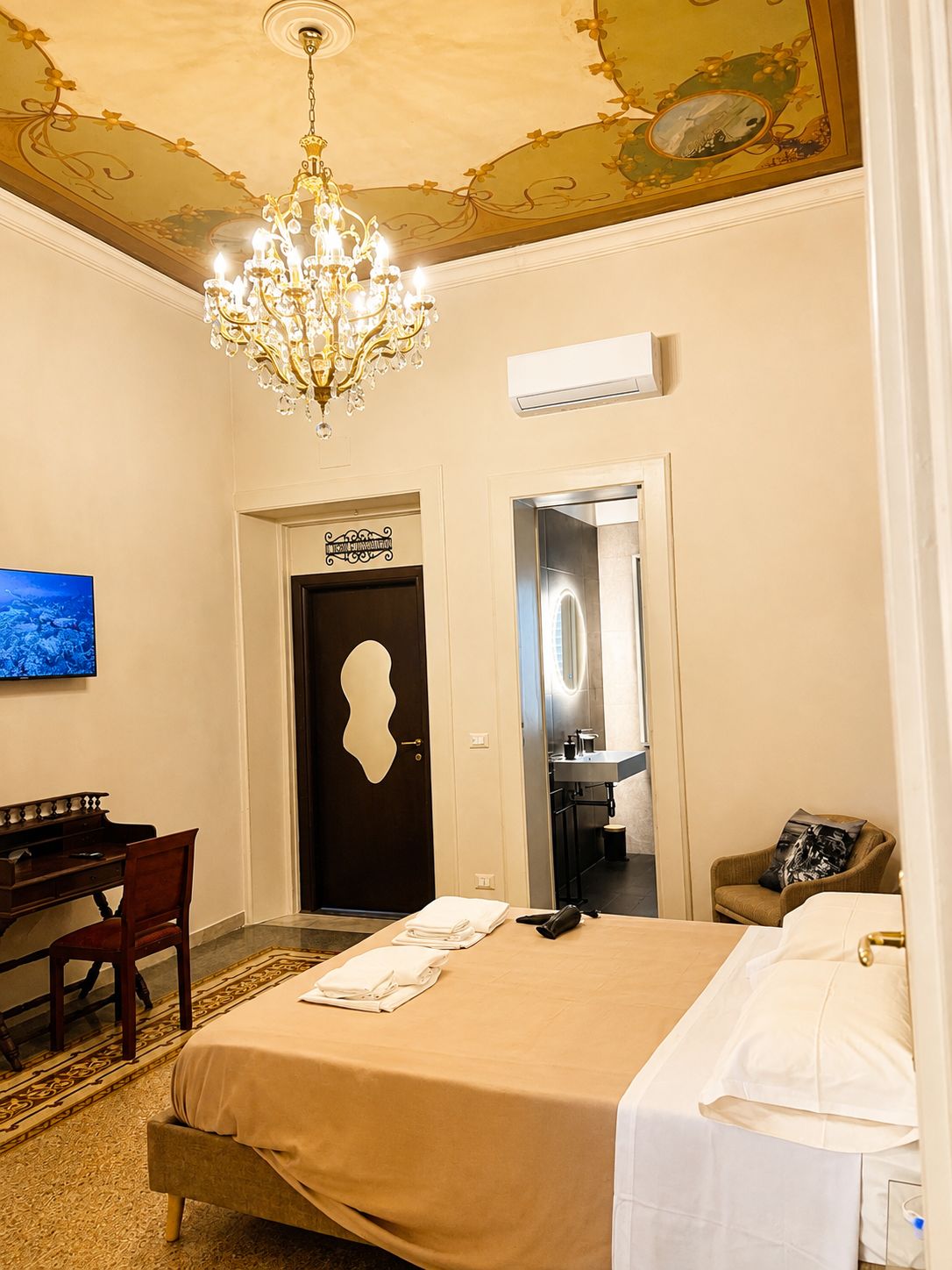 A hotel room with a bed, a desk, a chair, a television, a chandelier, and a bathroom visible through an open door.