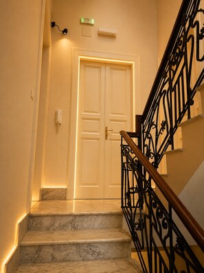 A marble staircase with black wrought iron railings leading to a double door with a light switch above it. - The Noble Stay (Gallico)