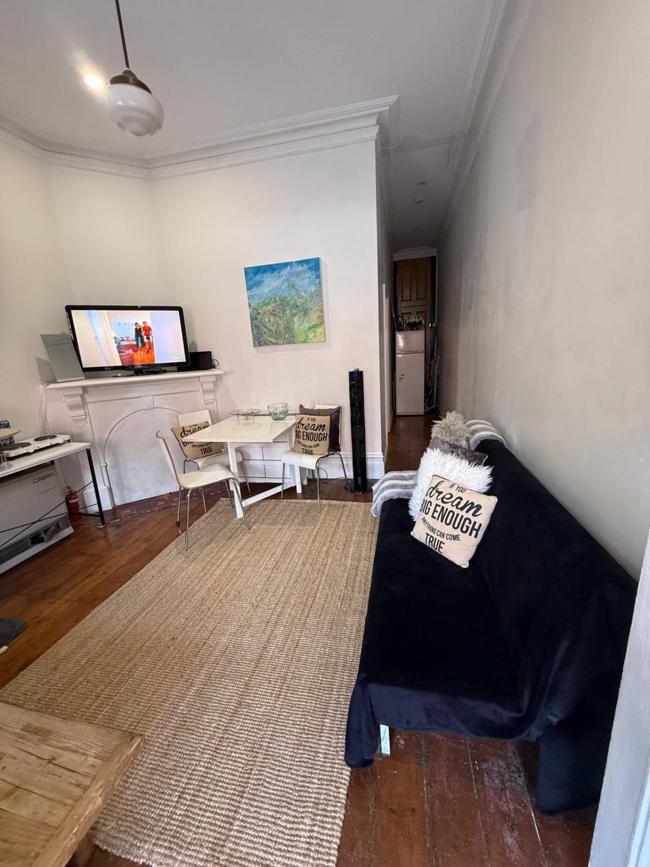 Uma sala de estar com um sofá preto, uma mesa de centro, uma TV em um suporte e um quadro na parede.