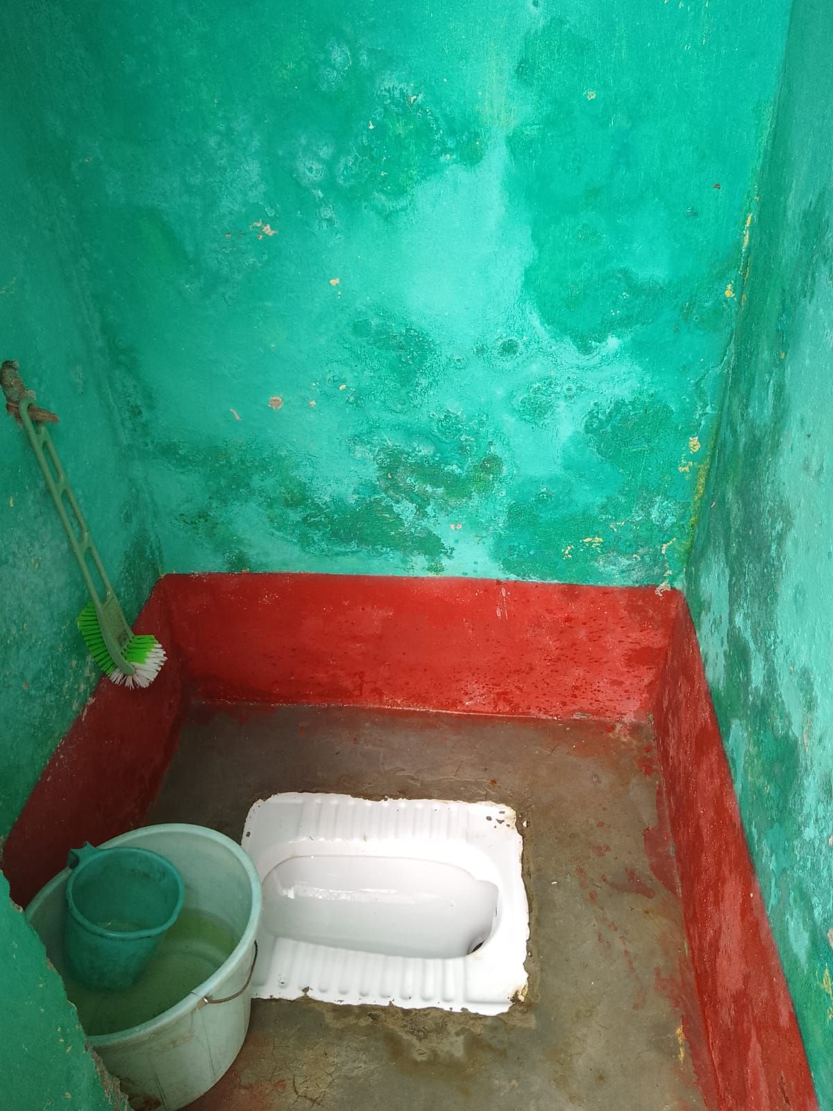 Een kleine, smalle ruimte met een enkel wit toilet, een groene emmer en een bezem.