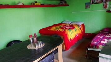 A room with a bed, a table, and a chair. There is a shelf above the bed and a wall with pictures.