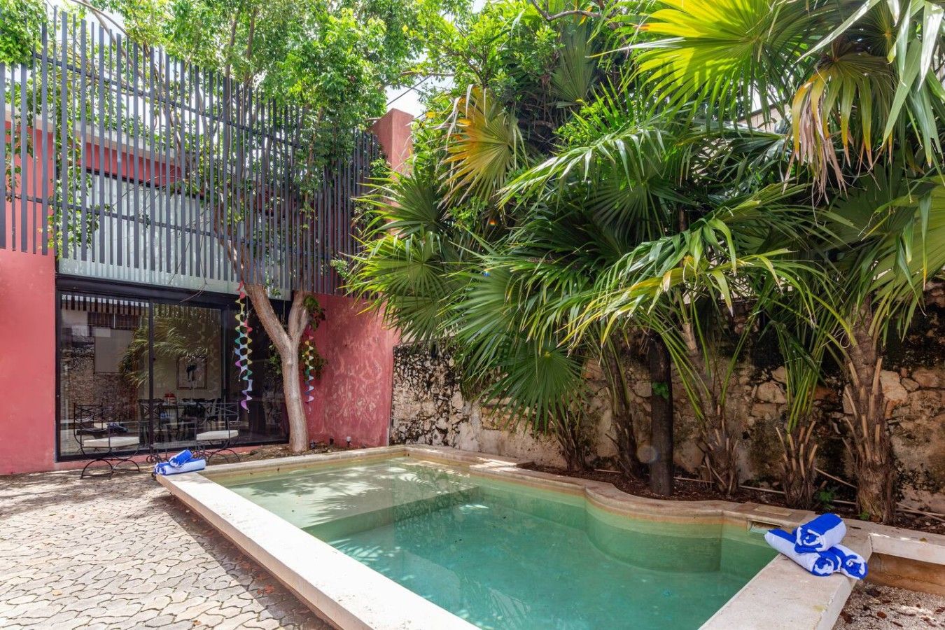 Una piscina con borde de piedra, rodeada de exuberante vegetación y un edificio rojo con puerta de cristal.