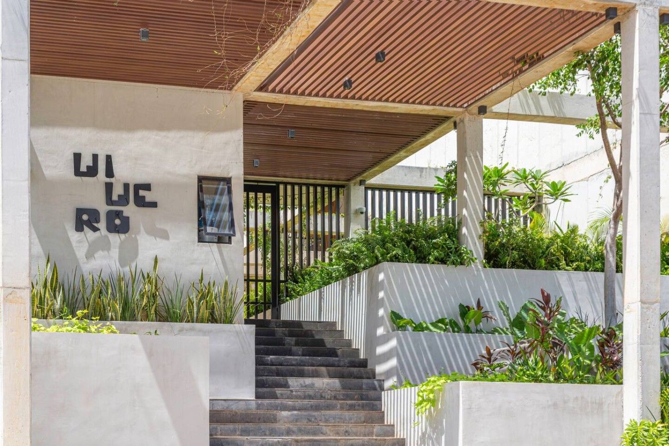 A modern building with a concrete facade, a wooden slatted roof, and a staircase leading to a gated entrance.
