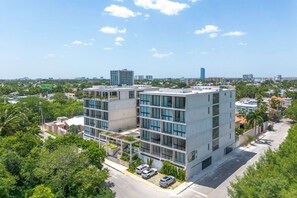 Um edifício moderno de vários andares com telhado plano, rodeado por vegetação e outros edifícios.