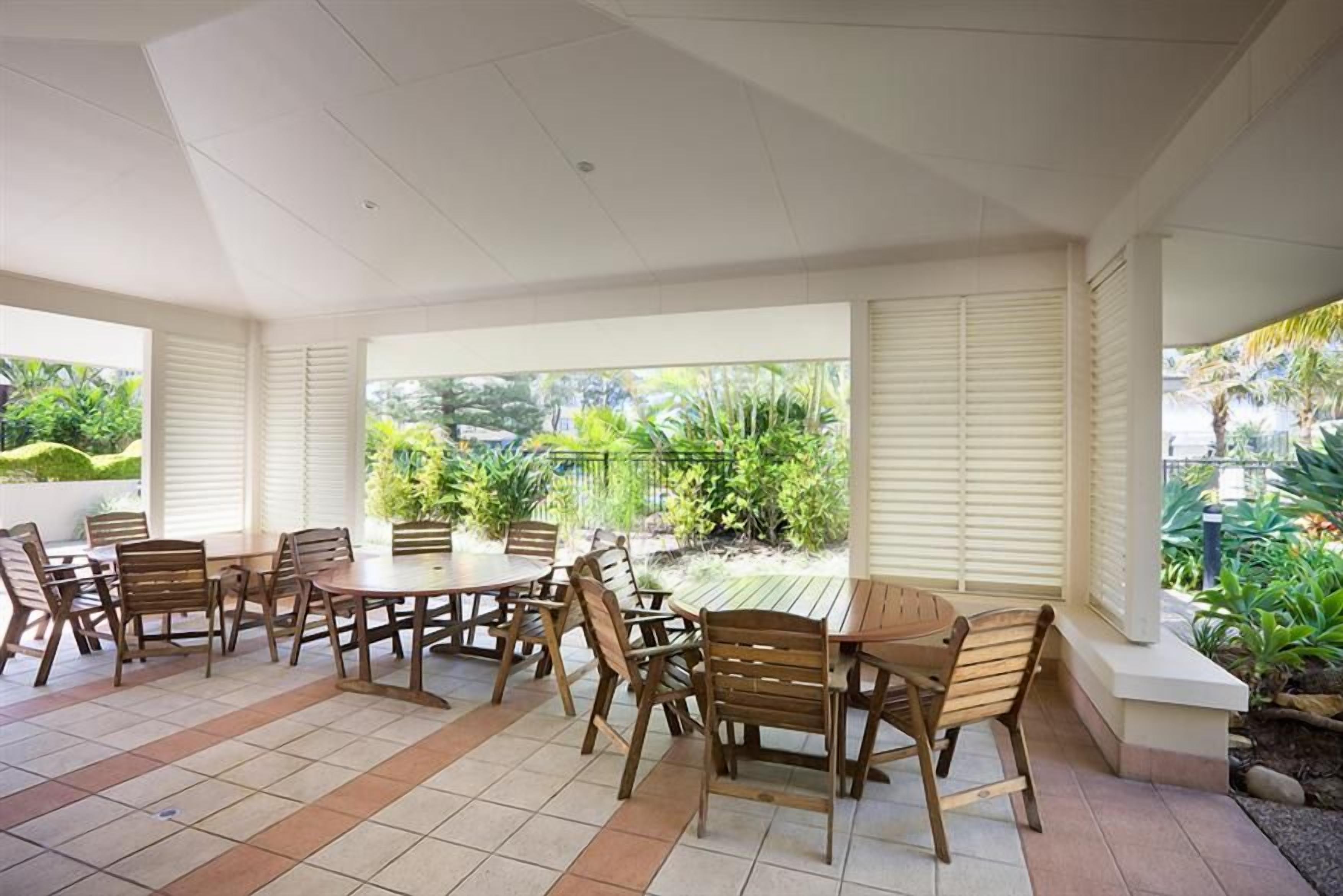 A covered outdoor seating area with tables and chairs, surrounded by plants and a view of greenery.