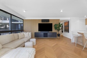 Ein modernes Wohnzimmer mit einer Couch, einem Flachbildfernseher, einer Essecke mit Hocker und einem großen Fenster mit Blick auf eine Stadtlandschaft.