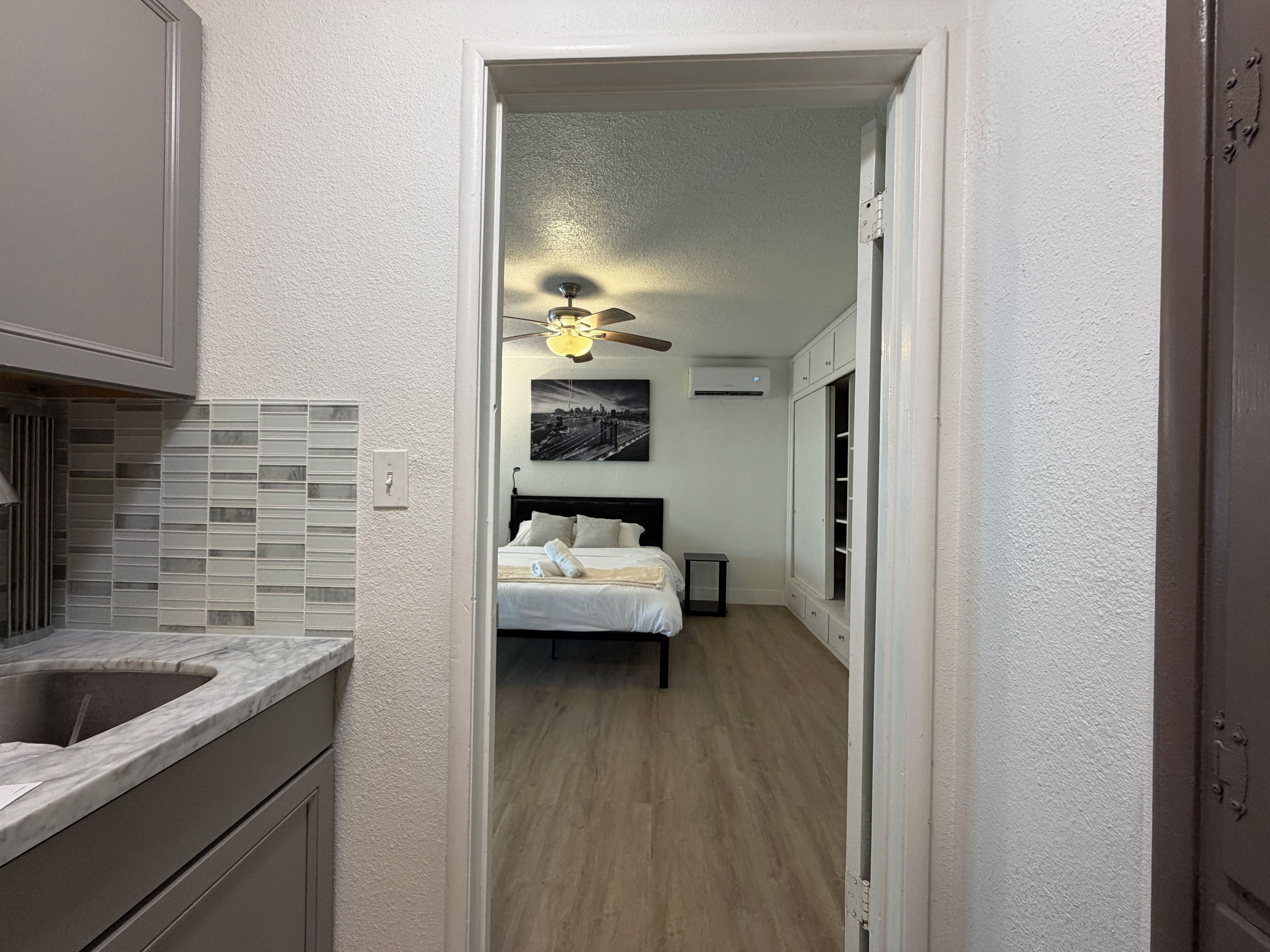 Un pasillo estrecho que conduce a un dormitorio con una cama, un ventilador de techo y un cuadro de un paisaje urbano.