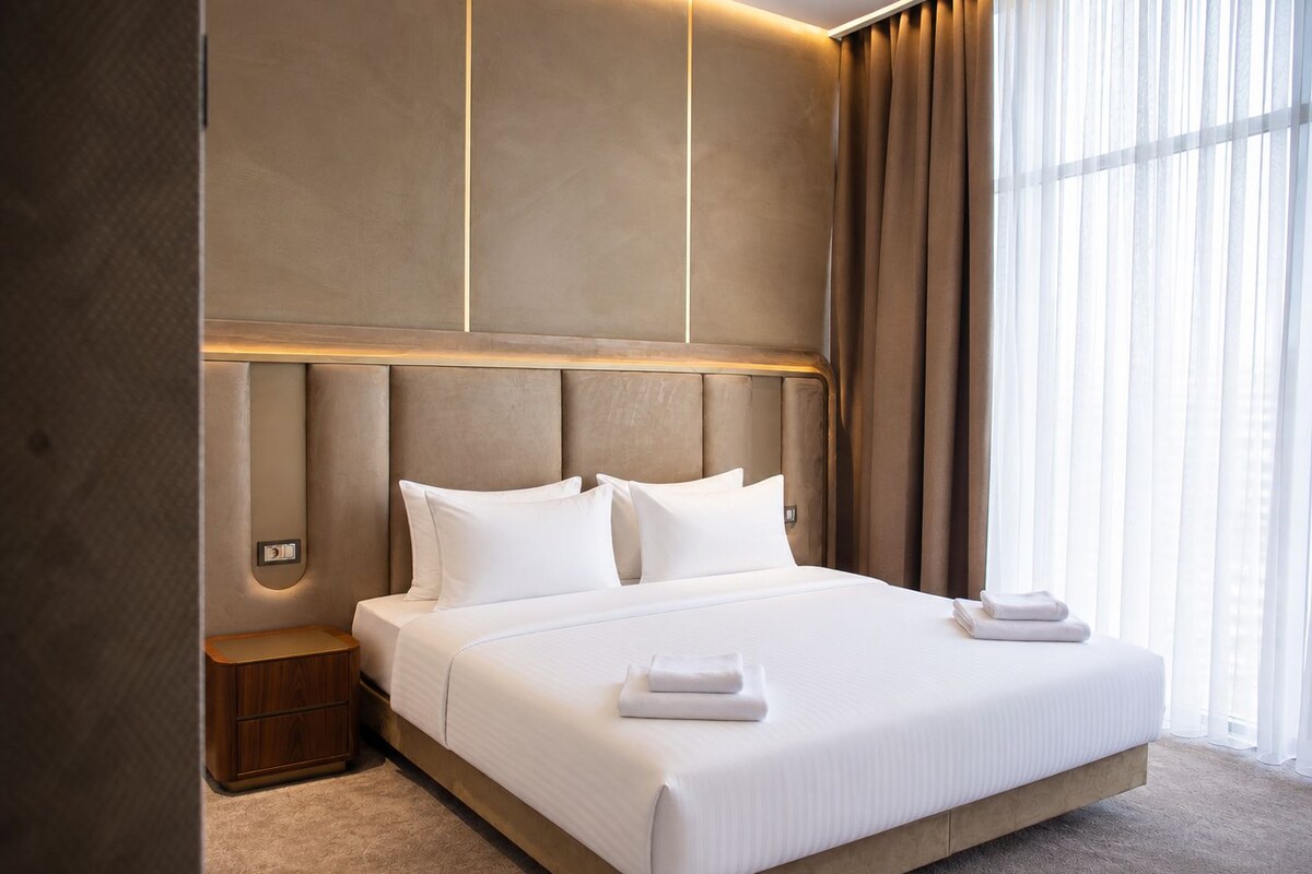 A modern hotel room with a large bed, a nightstand, and a window with curtains.