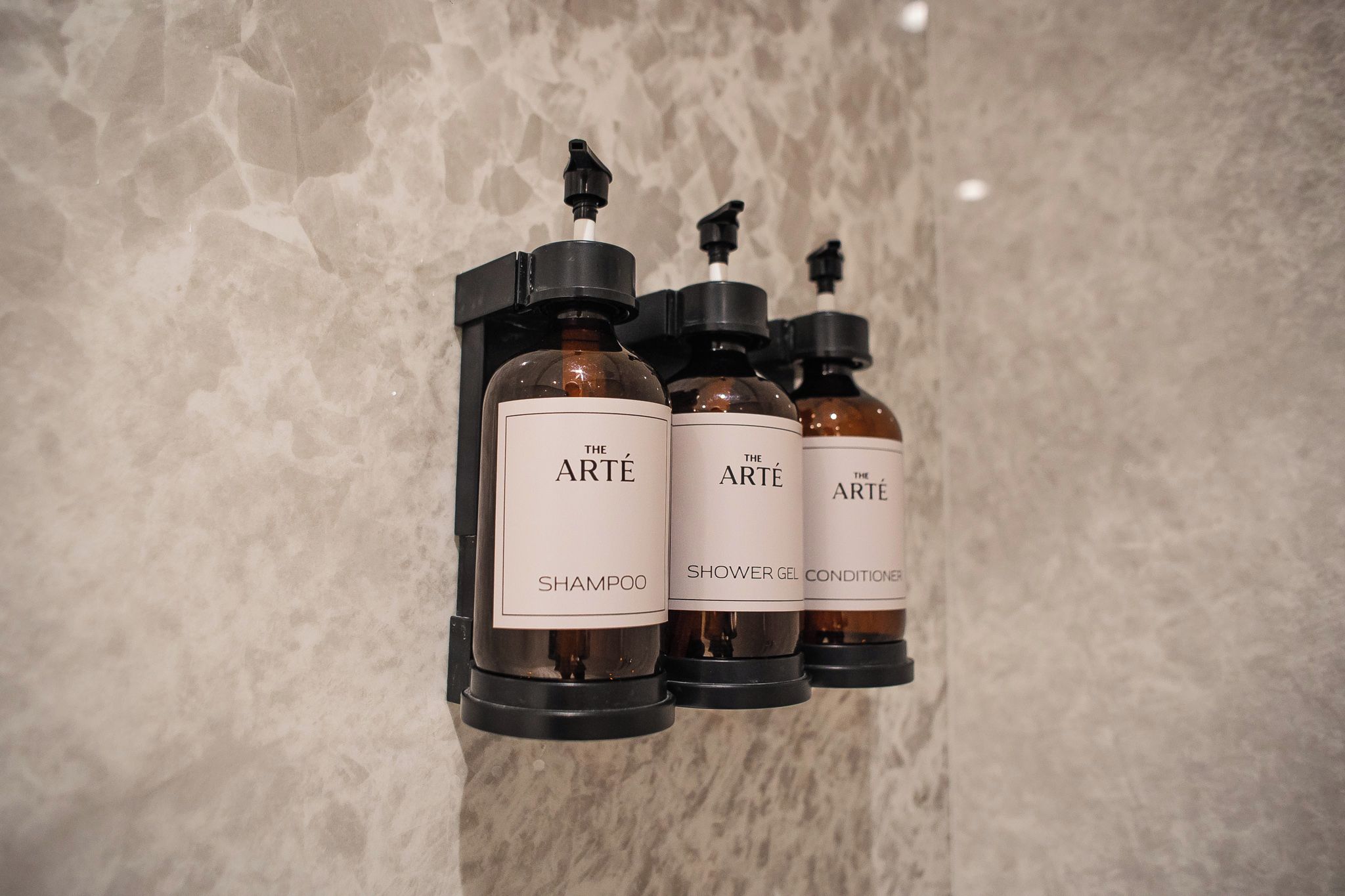Tres botellas de productos de la marca Arté: champú, gel de ducha y acondicionador, sujetas a una pared.