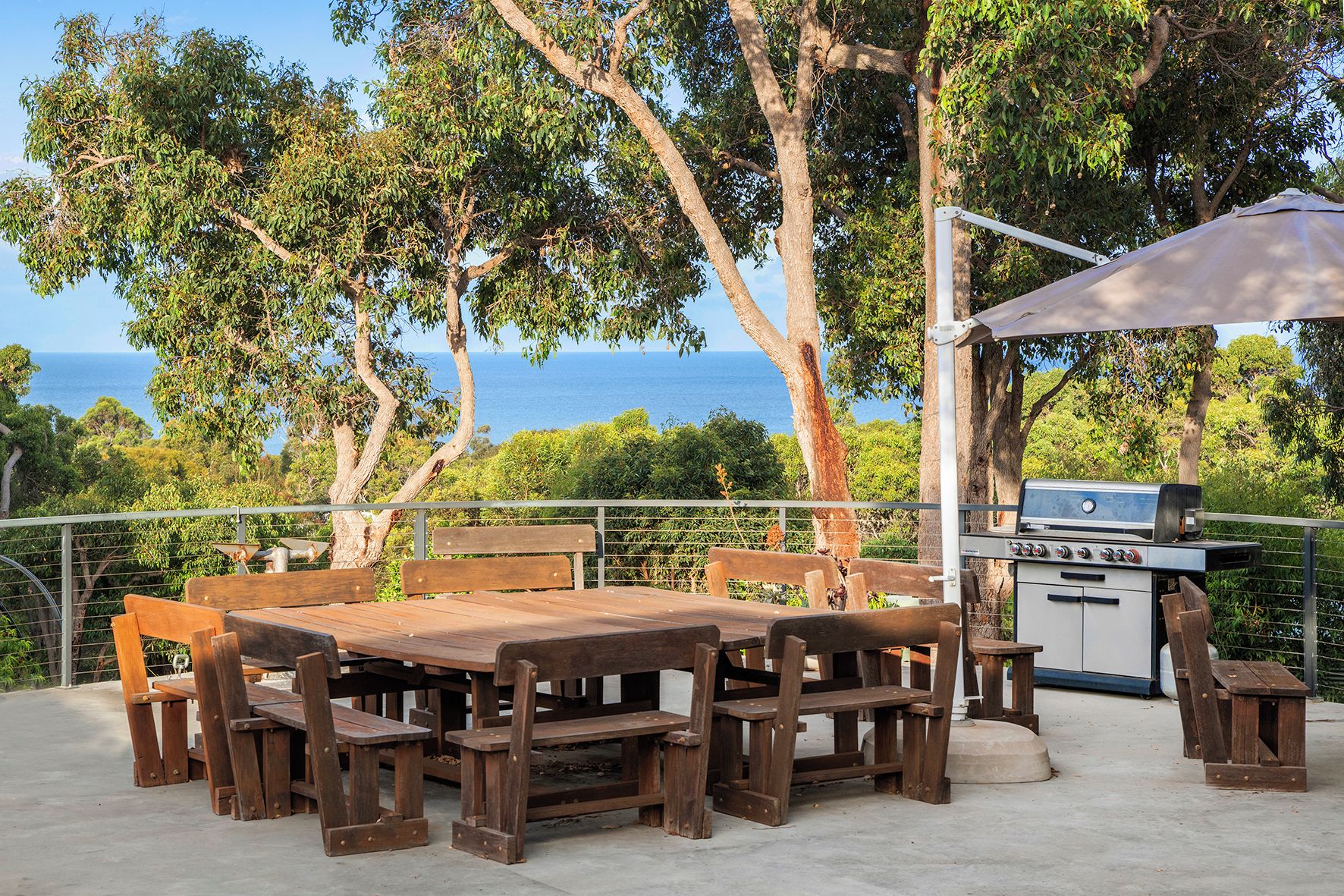 Patio esterno con tavolo e sedie in legno, barbecue e vista sul mare.