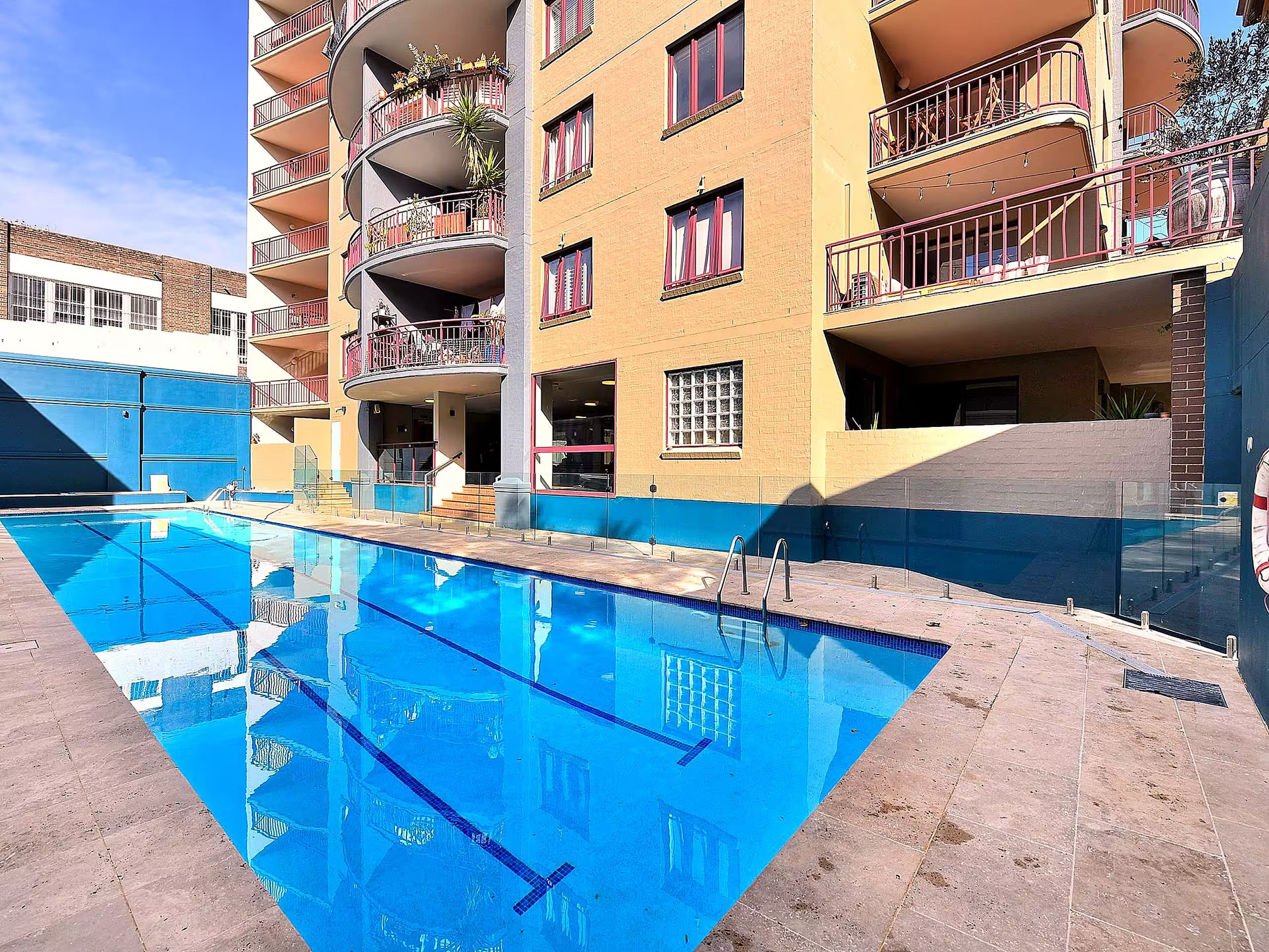 Una piscina con superficie de agua azul cristalina, rodeada por un edificio de varios pisos con balcones y ventanas.