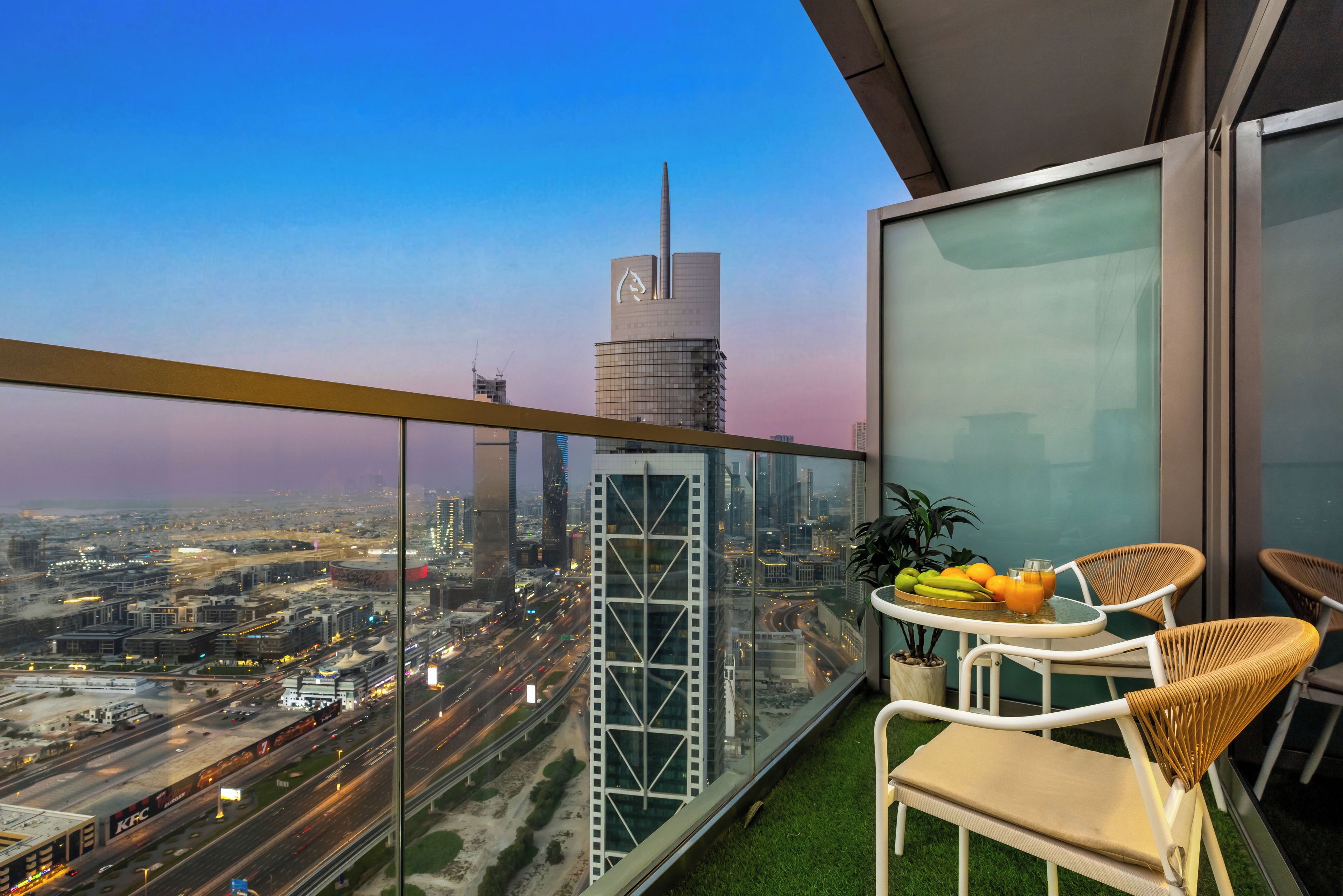 Een balkon met uitzicht op een stadsgezicht, inclusief een hoog gebouw met een opvallend logo, een tafel met fruit en een glas sap, en twee rieten stoelen.