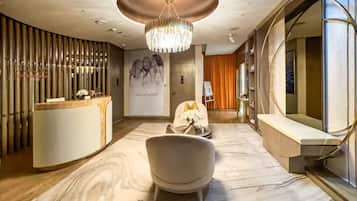 En modern hotellobby med en receptionsdisk, en stor kristallkrona, en sittgrupp med en fåtölj och en vas med blommor samt en stor spegel.