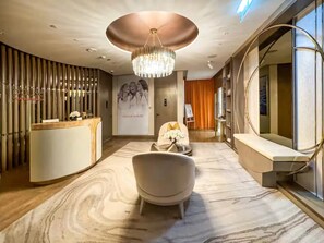 Un moderno vestíbulo de hotel con un mostrador de recepción, una gran lámpara de araña, una zona de estar con una silla y un jarrón de flores, y un espejo grande.