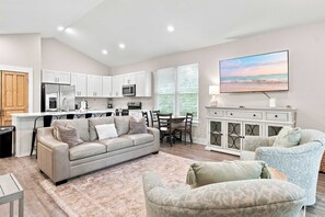 A modern kitchen with white cabinets, a refrigerator, and a microwave. There is a dining area with a table and chairs, and a living room with a sofa and a TV. - Coastal Getaway in Perdido Key (Pensacola)