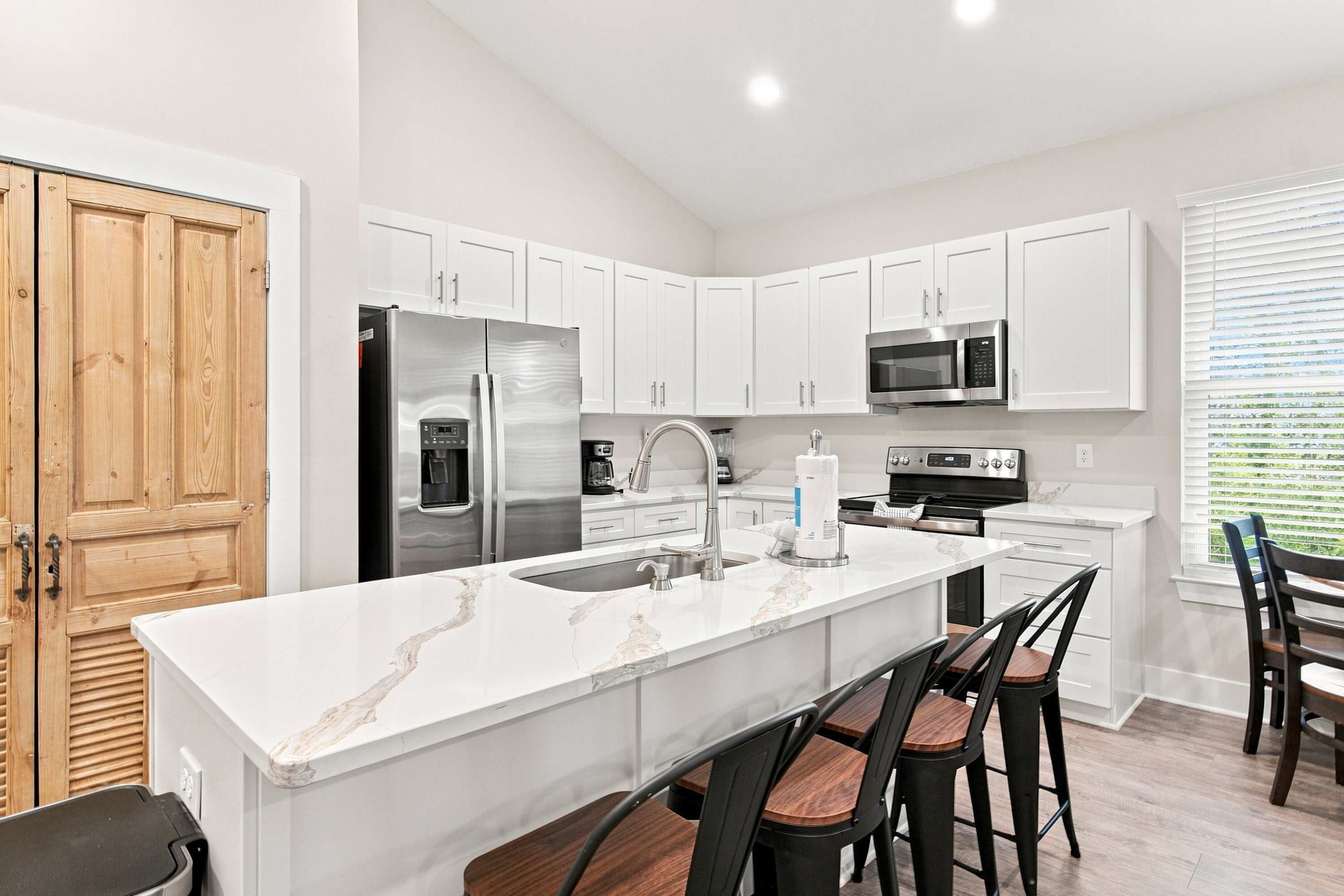A modern kitchen with white cabinetry, stainless steel appliances, a marble countertop, and a dining area with bar stools.