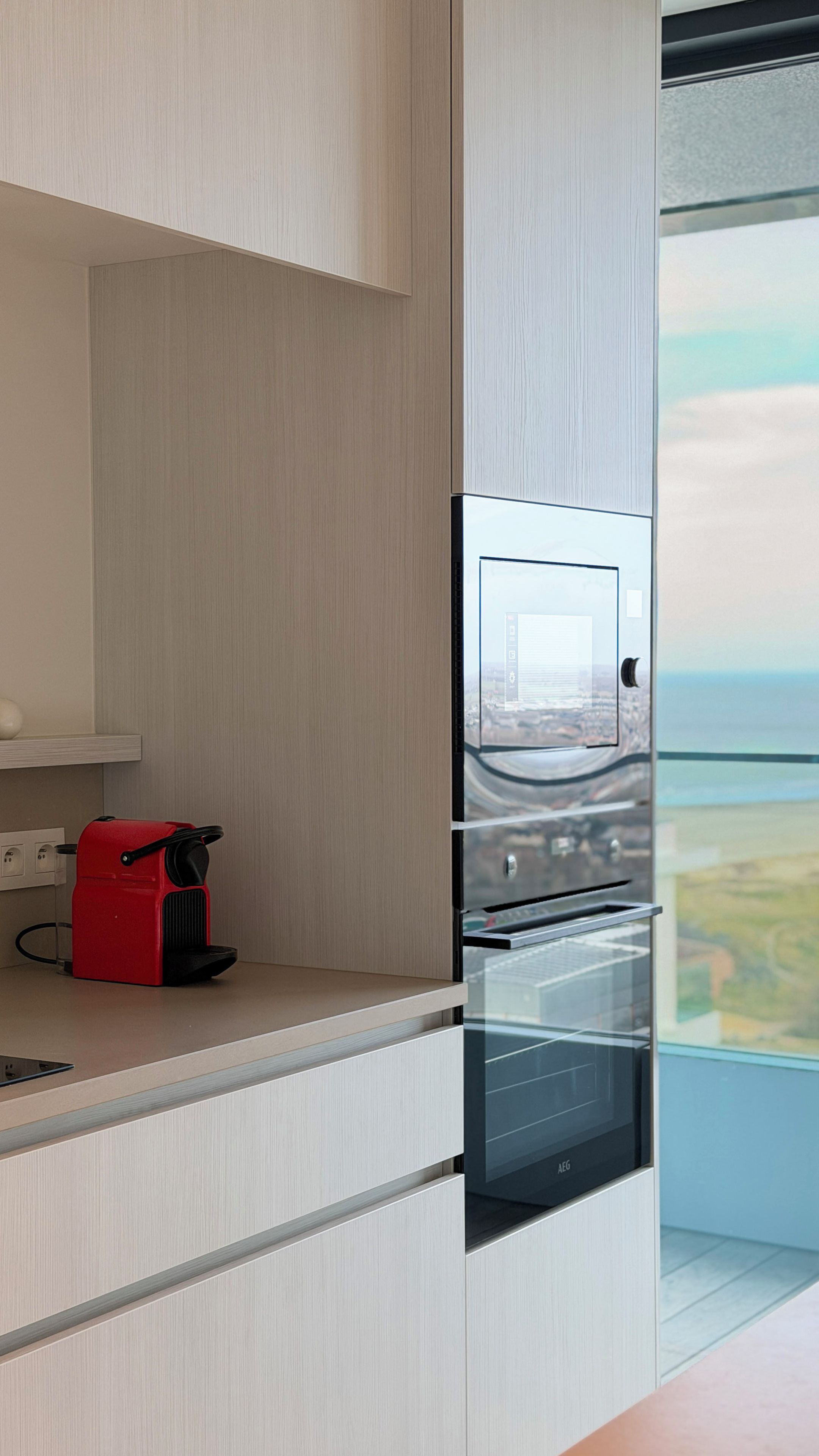 Una cocina moderna con electrodomésticos integrados, un hervidor rojo y vistas de la ciudad a través de un amplio ventanal.
