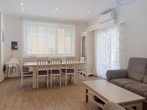 Een woonkamer met een eettafel, stoelen, een bank en een raam met jaloezieën.