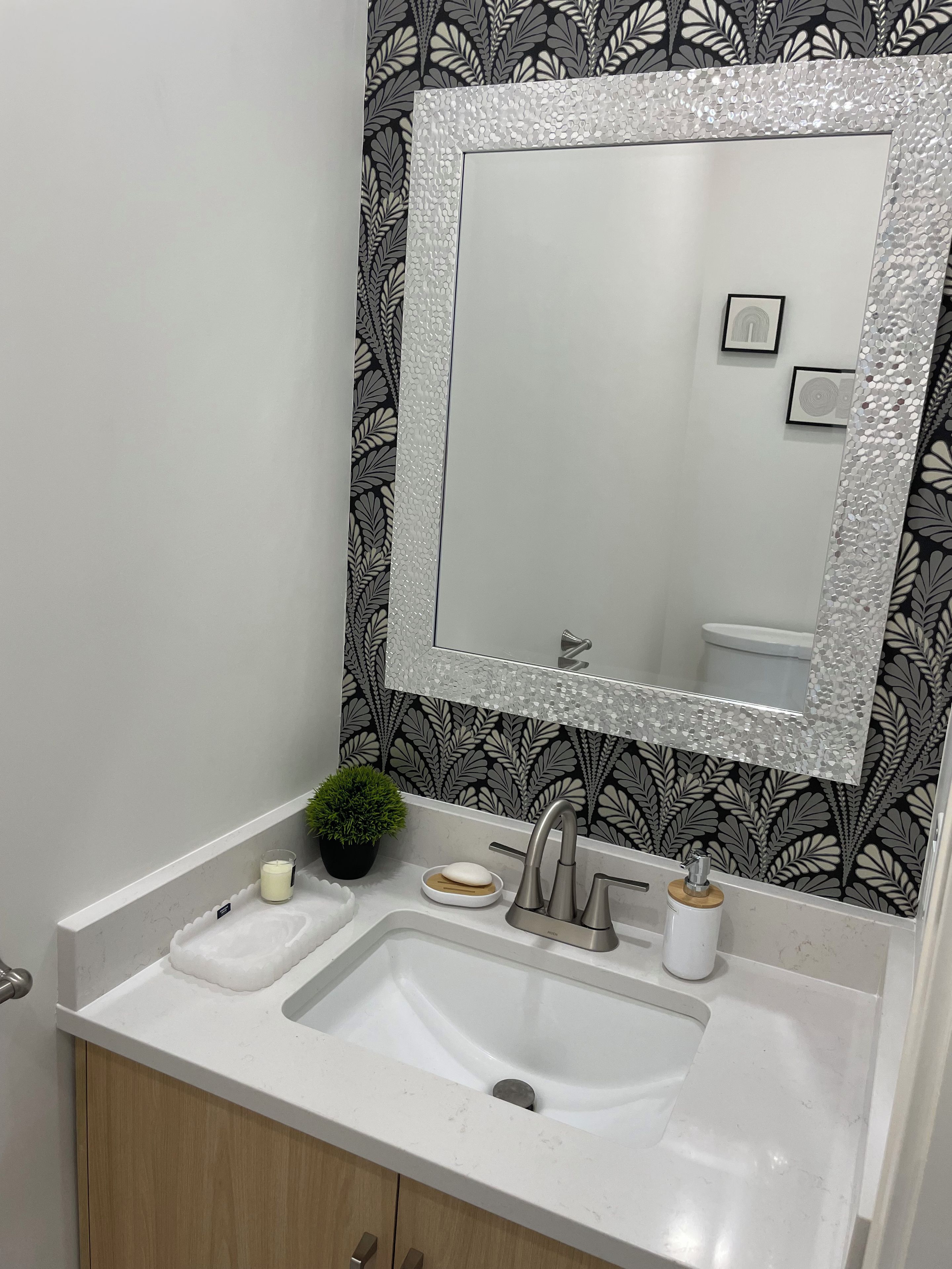 Een badkamer met een wit wastafelmeubel, een spiegel met een reliëf lijst, een plant in pot en een behang met dessin.