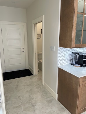 A hallway leading to a bathroom with a toilet and sink, a white door with a doormat, and a kitchen area with a coffee maker and a tiled backsplash. - Beautiful modern home in a quiet neighbourhood in Charlottetown (Charlottetown)