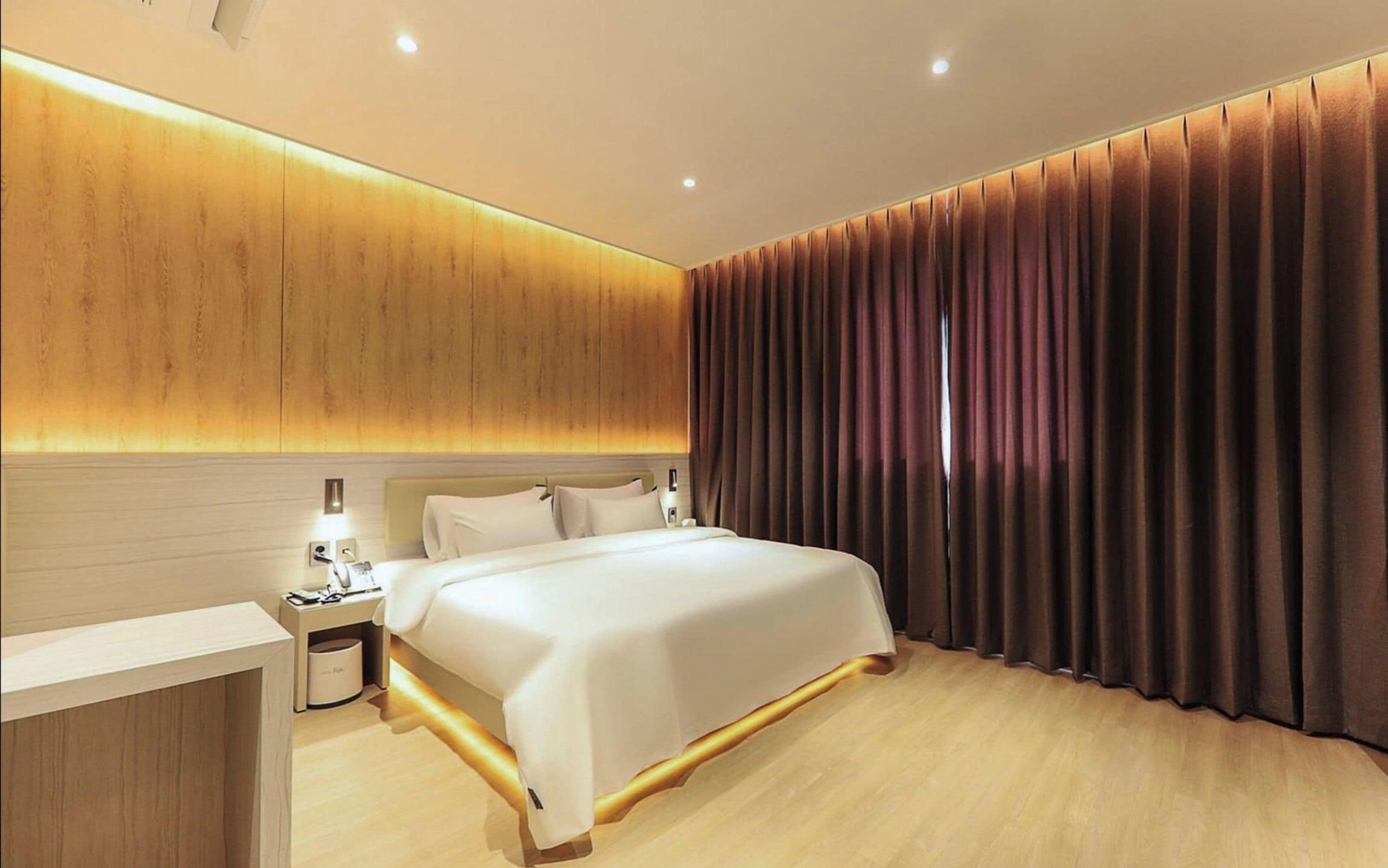 Habitación de hotel moderna con una cama grande, mesitas de noche, un cabecero de madera y vistas de la ciudad a través de cortinas.