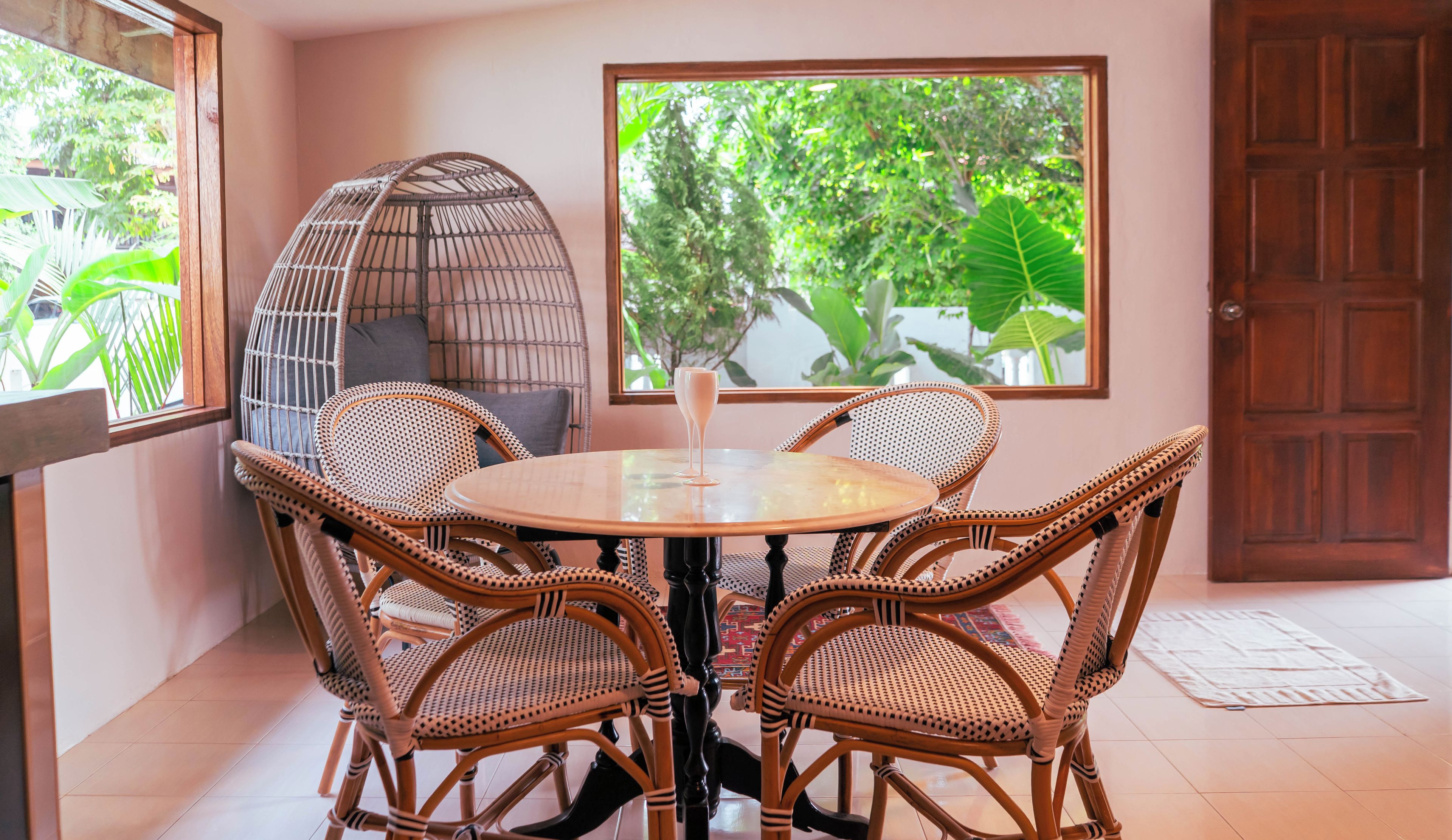 Una habitación con una mesa redonda, cuatro sillas de mimbre, un banco acolchado y un ventanal con vistas al verdor.