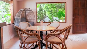 Una habitación con una mesa redonda, cuatro sillas de mimbre, un banco acolchado y un ventanal con vistas al verdor.
