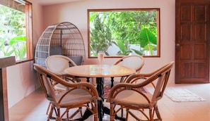 Una habitación con una mesa redonda, cuatro sillas de mimbre, un banco acolchado y un ventanal con vistas al verdor.