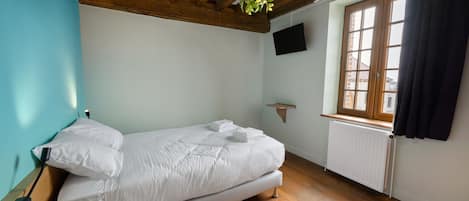 Une chambre à coucher avec un lit, un téléviseur fixé au mur, une fenêtre avec des rideaux et une plante suspendue.
