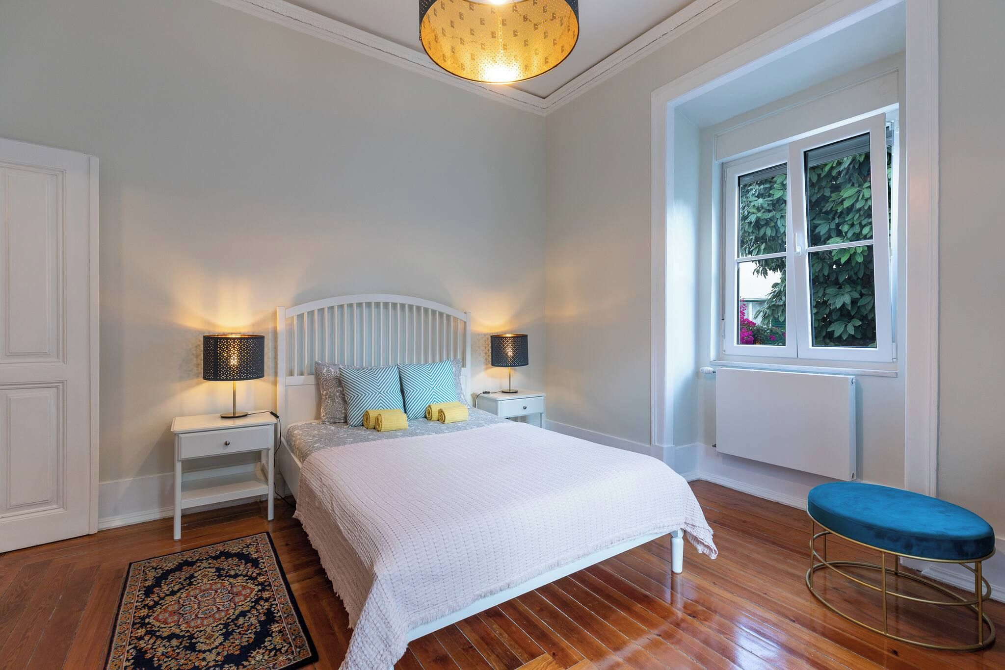 Une chambre à coucher avec un lit, deux tables de chevet avec des lampes, une fenêtre et un banc bleu.