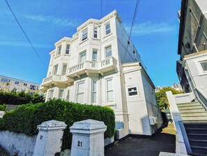 A multi-story building with a white facade, a balcony on the second floor, and a clear sky. - A restored Victorian apartment blending period elegance with modern comfort. (Hastings)