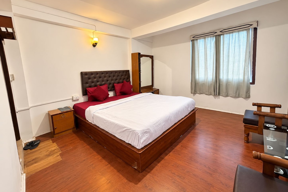 A bedroom with a wooden bed, red pillows, a nightstand, a mirror, a window with curtains, and a seating area with a table and chairs.