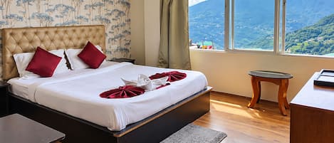 Habitación de hotel con una cama grande, una mesita de noche de madera, una pequeña mesa y vistas a un paisaje montañoso.