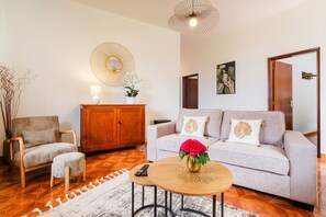 A living room with a sofa, armchair, wooden cabinet, and a round coffee table with a vase of flowers. - GuestReady - Quinta A Lealdade (Arco de São Jorge)